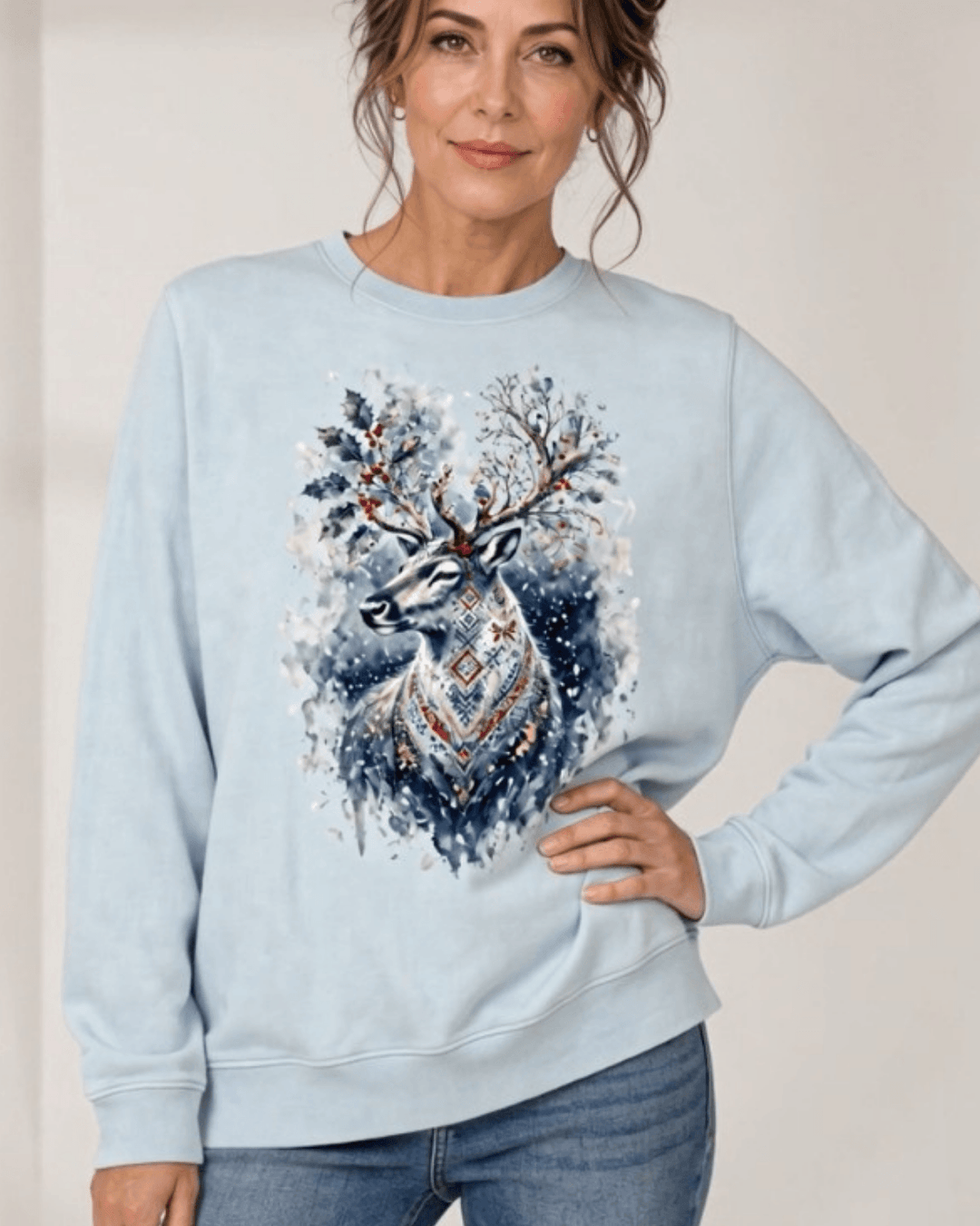 Made in USA Reindeer in the Snow Christmas Design crew neck sweatshirt in light blue with a watercolor-style reindeer graphic with holly in its antlers and snowy splatters, front view with hand on hip on female model with jeans