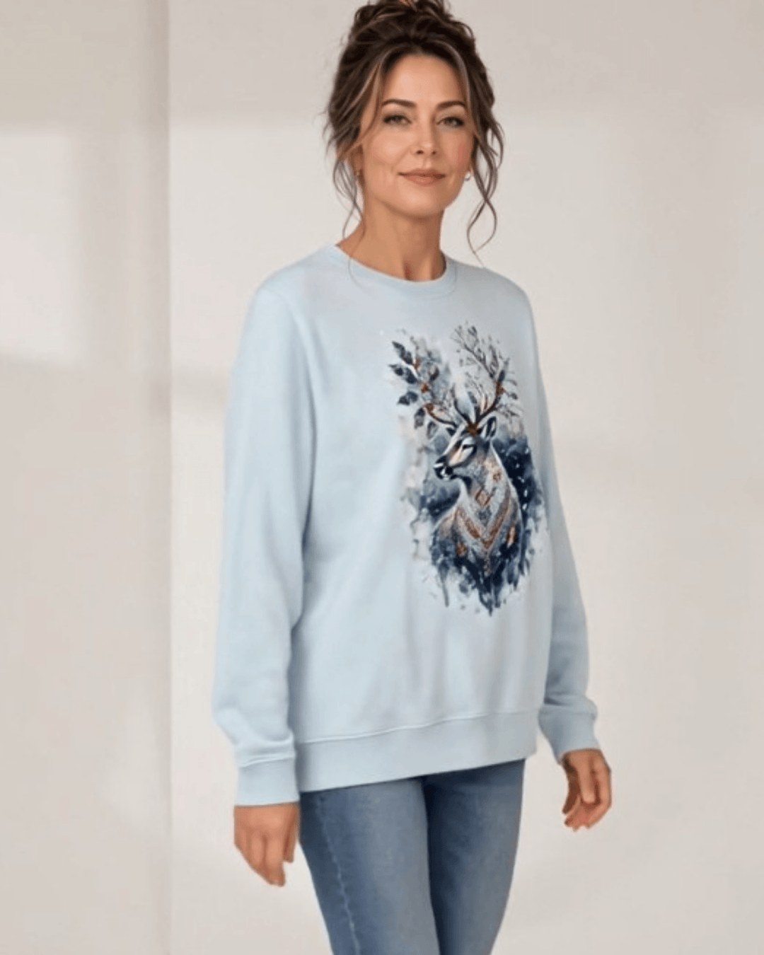 Made in USA Reindeer in the Snow Christmas Design crew neck sweatshirt in light blue with a watercolor-style reindeer graphic with holly in its antlers and snowy splatters, front full-length walking view on female model with jeans