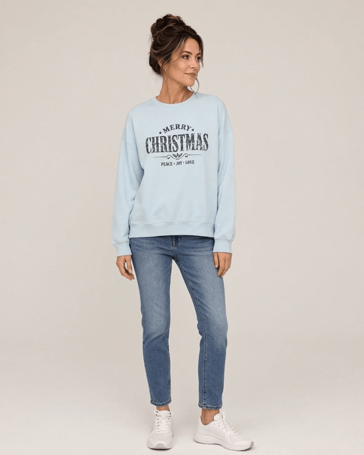 Made in USA Vintage Distressed Merry Christmas crew neck sweatshirt in light blue with a distressed charcoal MERRY CHRISTMAS graphic with PEACE JOY LOVE, front full-length view looking to the side on female model with jeans