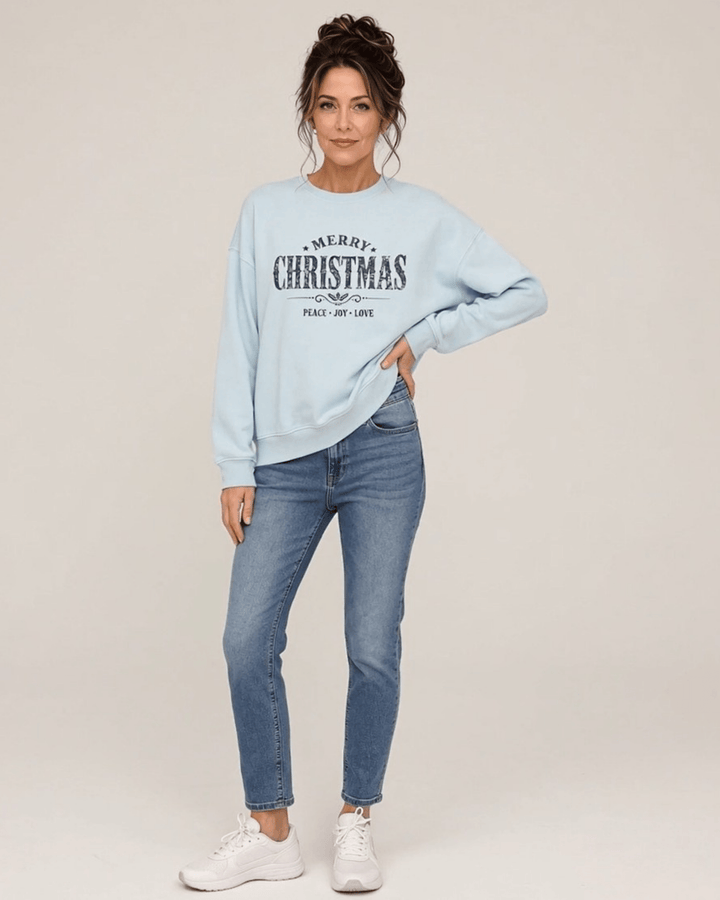 Made in USA Vintage Distressed Merry Christmas crew neck sweatshirt in light blue with a distressed charcoal MERRY CHRISTMAS graphic with PEACE JOY LOVE, front full-length view with hand on hip on female model with jeans