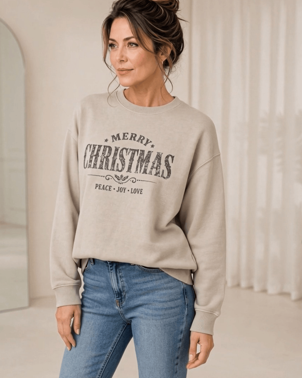 Made in USA Vintage Distressed Merry Christmas crew neck sweatshirt in taupe with a distressed charcoal MERRY CHRISTMAS graphic with PEACE JOY LOVE, three-quarter front view looking to the side on female model with jeans